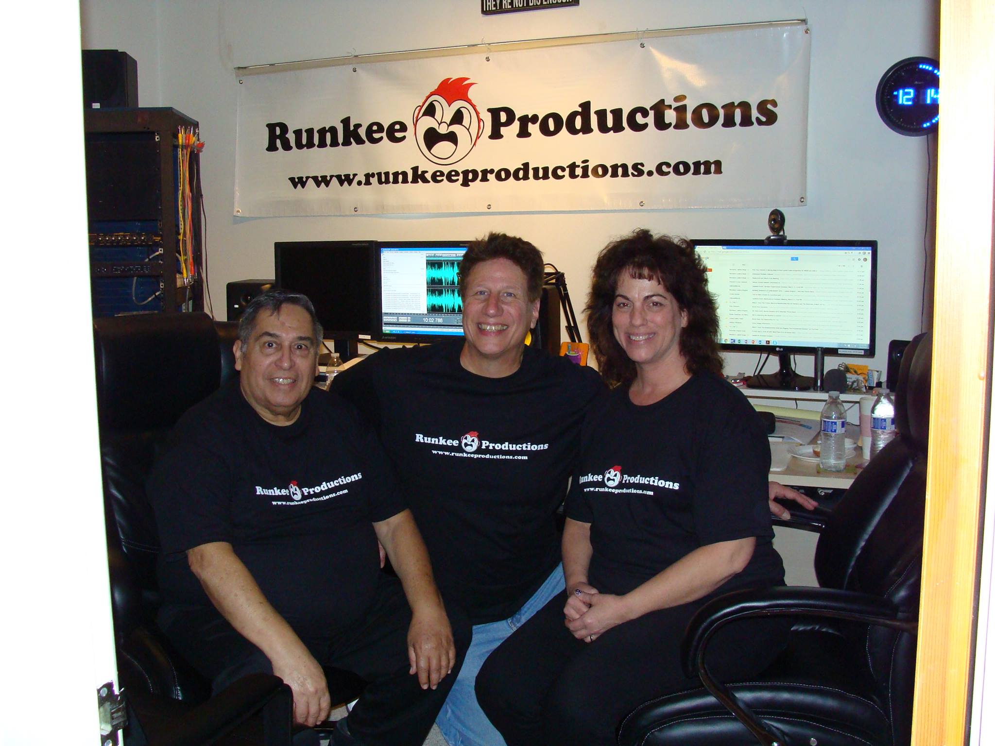 Three people pose in a studio setting, wearing matching black shirts featuring "Runkee Productions." Computers and audio equipment are visible in the background.